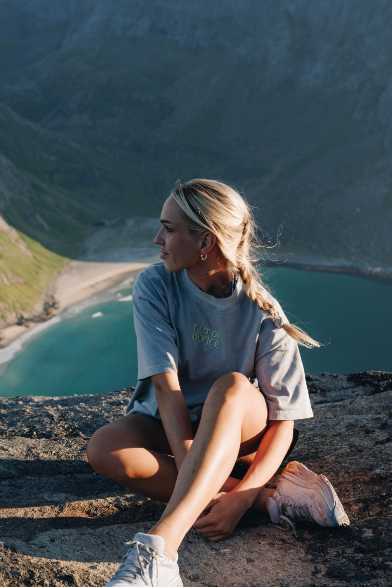 Kvinne som sitter opp på en fjellkant med en grå blå t skjorte med lime grønn logo midt på brystet